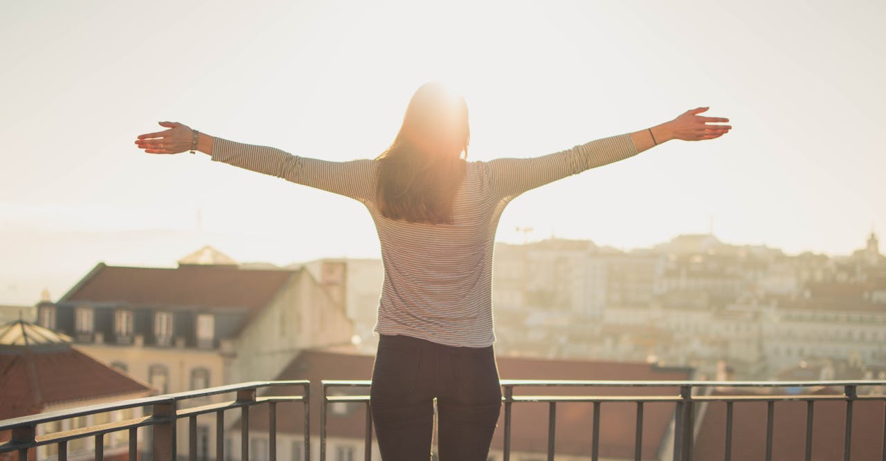 hero-img-02 A woman stands with outstretched arms on a sunny balcony, embracing the morning light.